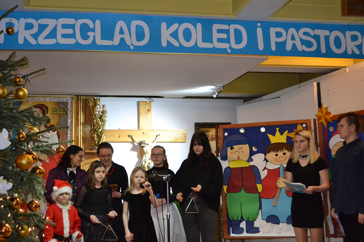 Dziewczynka w czarnej sukience trzyma mikrofon. Za śpiewającą pod ścianą stoją uczniowie, dwie dziewczynki trzymają instrumenty muzyczne - trójkąty.  