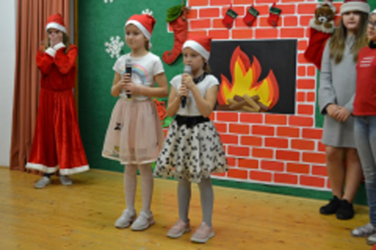 Uczniowie siedzą na krzesłach. Na głowach mają czerwone czapki Mikołaja. Oglądają występ na scenie. Mikołaj siedzi w fotelu na scenie i rozdaje prezenty.