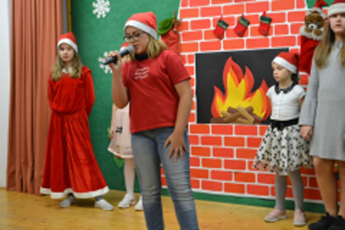 Uczniowie siedzą na krzesłach. Na głowach mają czerwone czapki Mikołaja. Oglądają występ na scenie. Mikołaj siedzi w fotelu na scenie i rozdaje prezenty.