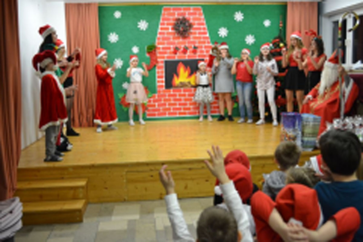 Uczniowie siedzą na krzesłach. Na głowach mają czerwone czapki Mikołaja. Oglądają występ na scenie. Mikołaj siedzi w fotelu na scenie i rozdaje prezenty.