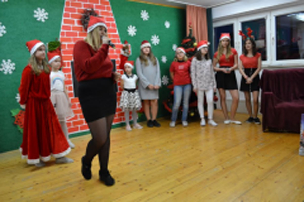 Uczniowie siedzą na krzesłach. Na głowach mają czerwone czapki Mikołaja. Oglądają występ na scenie. Mikołaj siedzi w fotelu na scenie i rozdaje prezenty.