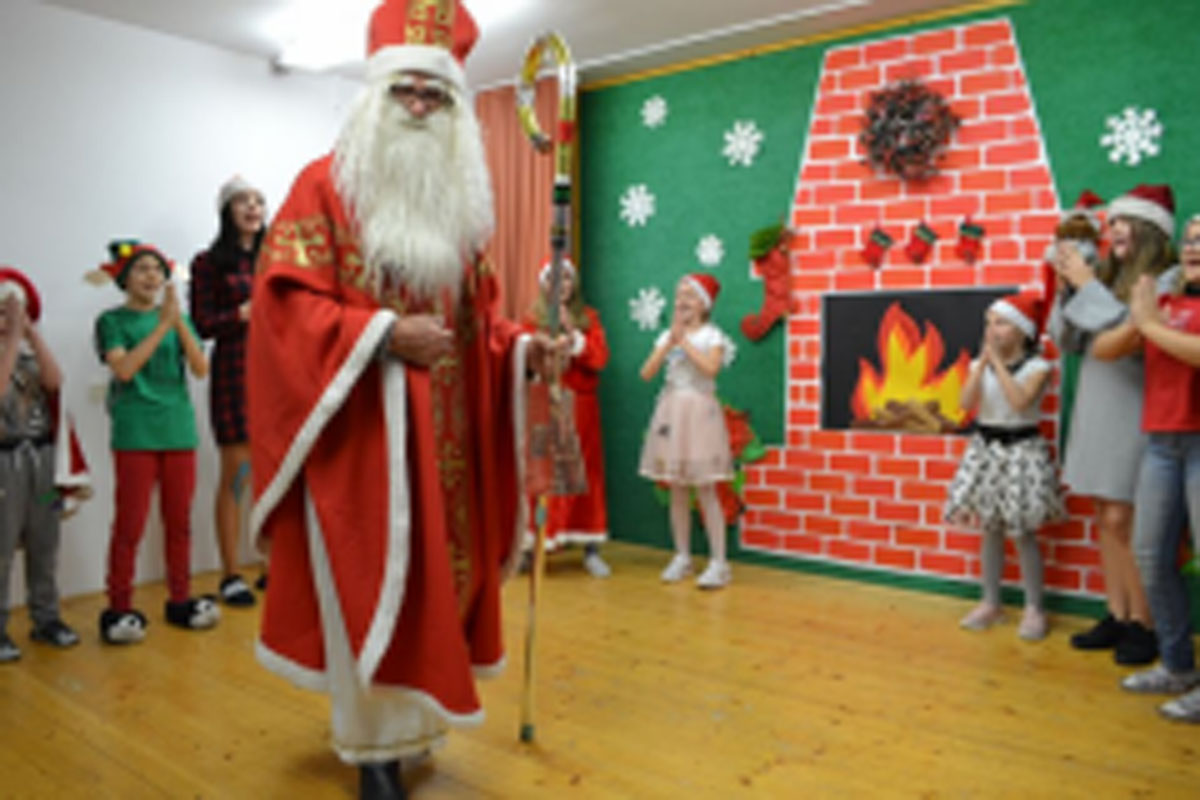 Uczniowie siedzą na krzesłach. Na głowach mają czerwone czapki Mikołaja. Oglądają występ na scenie. Mikołaj siedzi w fotelu na scenie i rozdaje prezenty.