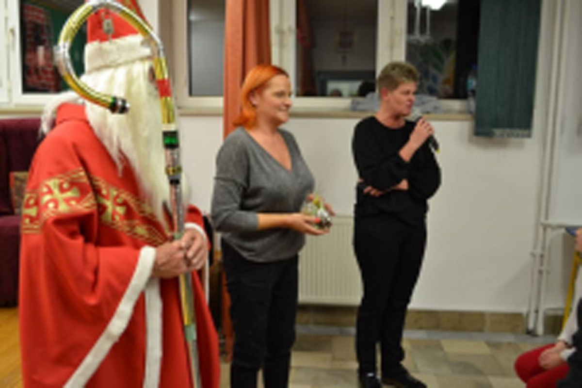 Uczniowie siedzą na krzesłach. Na głowach mają czerwone czapki Mikołaja. Oglądają występ na scenie. Mikołaj siedzi w fotelu na scenie i rozdaje prezenty.