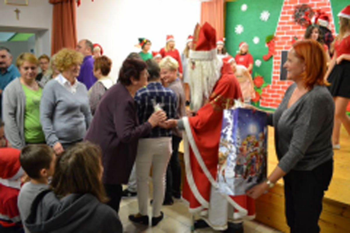 Uczniowie siedzą na krzesłach. Na głowach mają czerwone czapki Mikołaja. Oglądają występ na scenie. Mikołaj siedzi w fotelu na scenie i rozdaje prezenty.