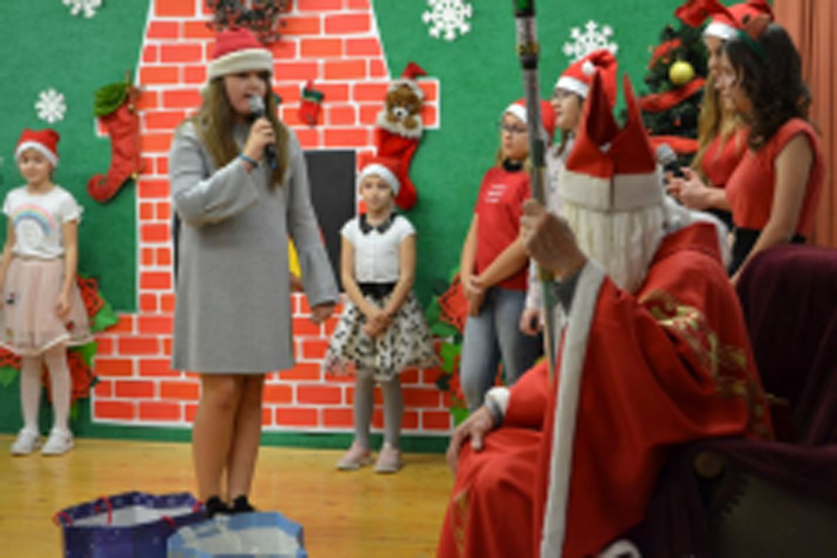 Uczniowie siedzą na krzesłach. Na głowach mają czerwone czapki Mikołaja. Oglądają występ na scenie. Mikołaj siedzi w fotelu na scenie i rozdaje prezenty.