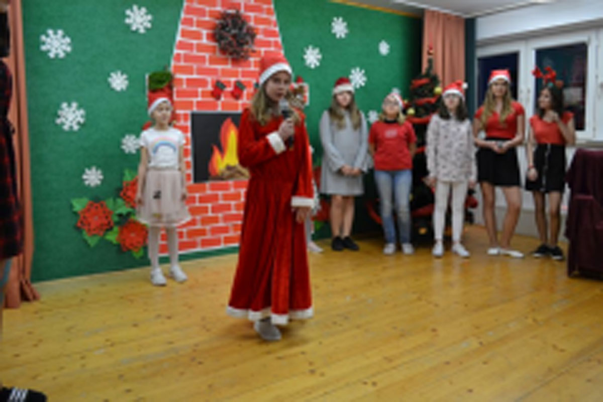 Uczniowie siedzą na krzesłach. Na głowach mają czerwone czapki Mikołaja. Oglądają występ na scenie. Mikołaj siedzi w fotelu na scenie i rozdaje prezenty.