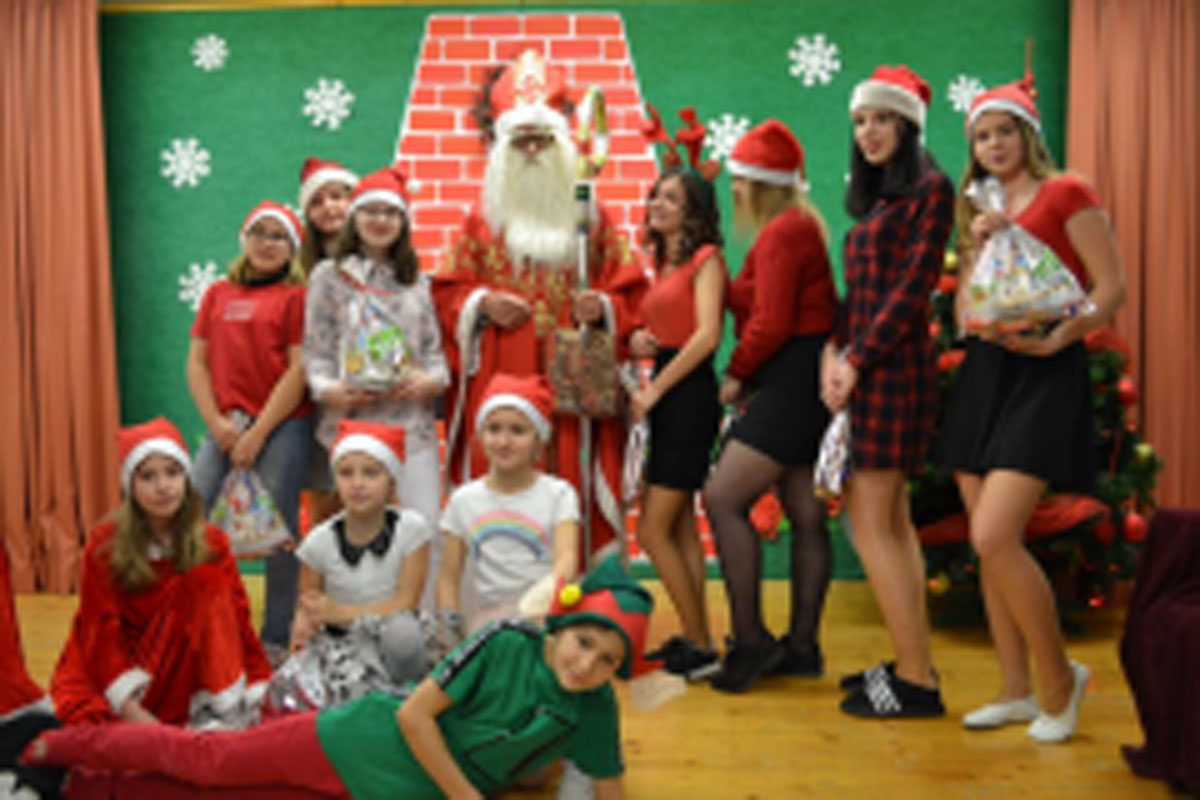 Uczniowie siedzą na krzesłach. Na głowach mają czerwone czapki Mikołaja. Oglądają występ na scenie. Mikołaj siedzi w fotelu na scenie i rozdaje prezenty.