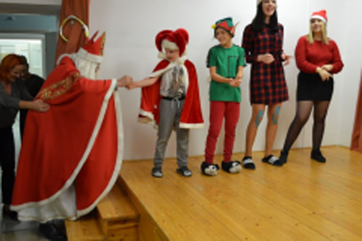 Uczniowie siedzą na krzesłach. Na głowach mają czerwone czapki Mikołaja. Oglądają występ na scenie. Mikołaj siedzi w fotelu na scenie i rozdaje prezenty.