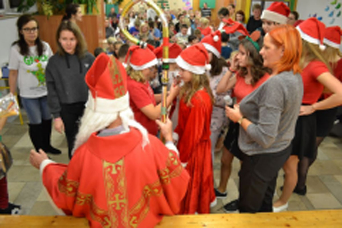 Uczniowie siedzą na krzesłach. Na głowach mają czerwone czapki Mikołaja. Oglądają występ na scenie. Mikołaj siedzi w fotelu na scenie i rozdaje prezenty.