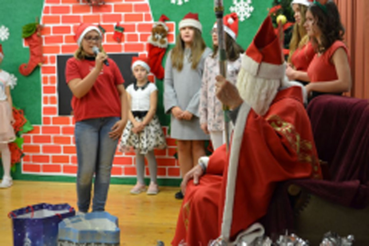 Uczniowie siedzą na krzesłach. Na głowach mają czerwone czapki Mikołaja. Oglądają występ na scenie. Mikołaj siedzi w fotelu na scenie i rozdaje prezenty.