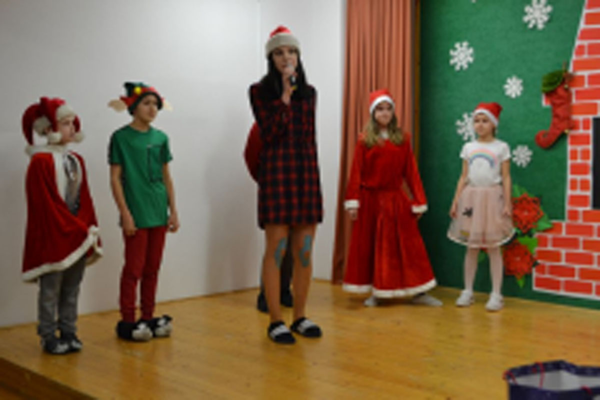 Uczniowie siedzą na krzesłach. Na głowach mają czerwone czapki Mikołaja. Oglądają występ na scenie. Mikołaj siedzi w fotelu na scenie i rozdaje prezenty.