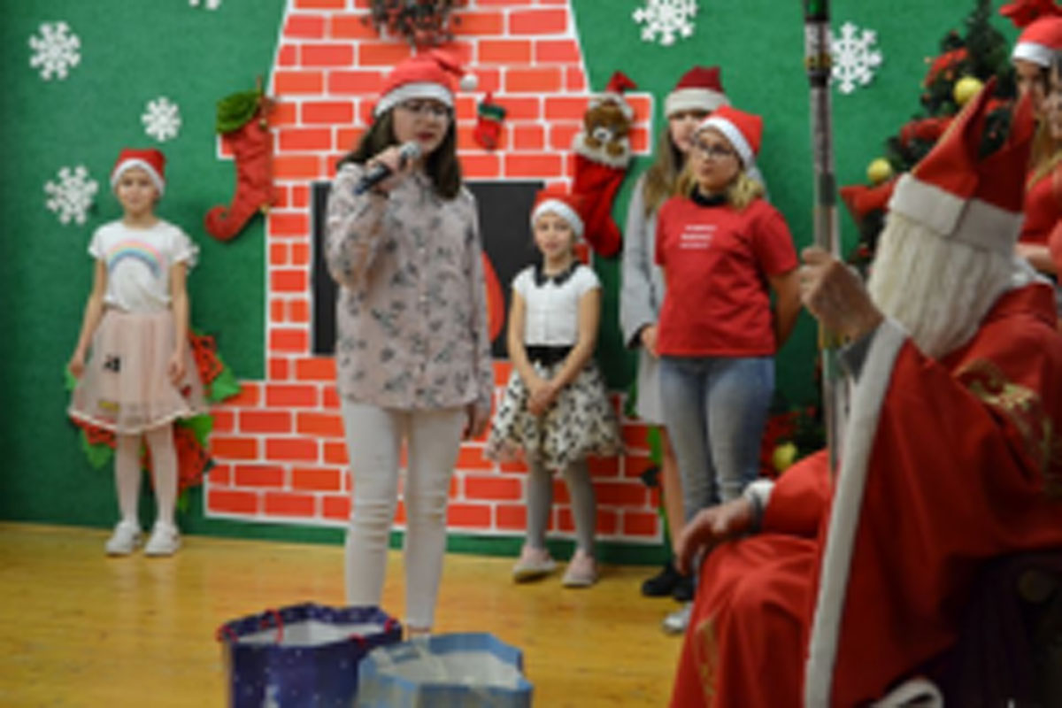 Uczniowie siedzą na krzesłach. Na głowach mają czerwone czapki Mikołaja. Oglądają występ na scenie. Mikołaj siedzi w fotelu na scenie i rozdaje prezenty.