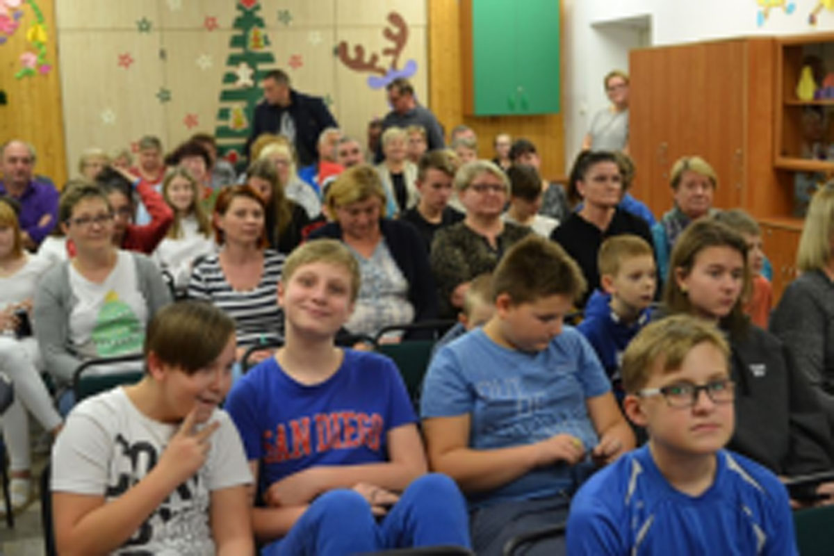 Uczniowie siedzą na krzesłach. Na głowach mają czerwone czapki Mikołaja. Oglądają występ na scenie. Mikołaj siedzi w fotelu na scenie i rozdaje prezenty.