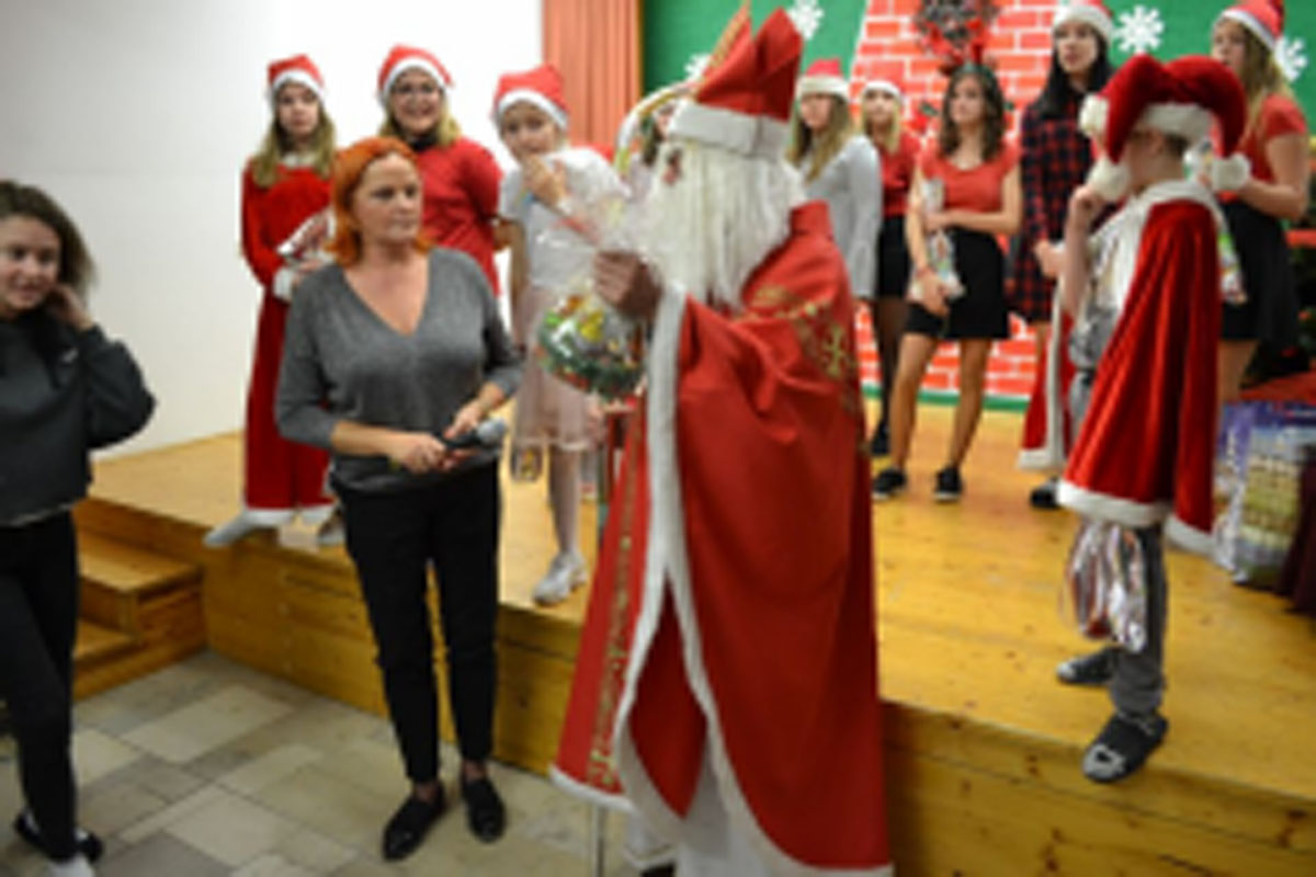 Uczniowie siedzą na krzesłach. Na głowach mają czerwone czapki Mikołaja. Oglądają występ na scenie. Mikołaj siedzi w fotelu na scenie i rozdaje prezenty.