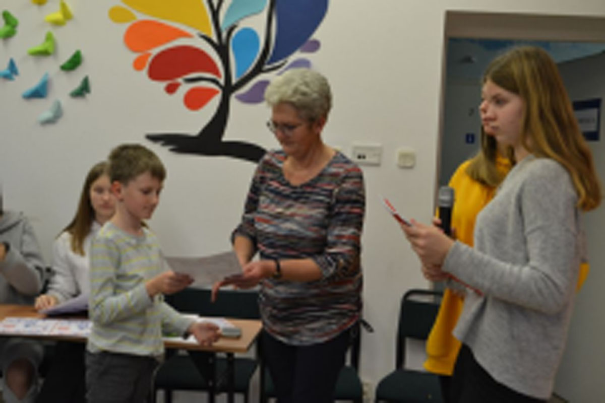 Uczniowie występują na scenie, trzymają mikrofony. Członkowie jury siedzą przy stoliku, wręczają dyplomy. Wspólne zdjęcie uczniów.