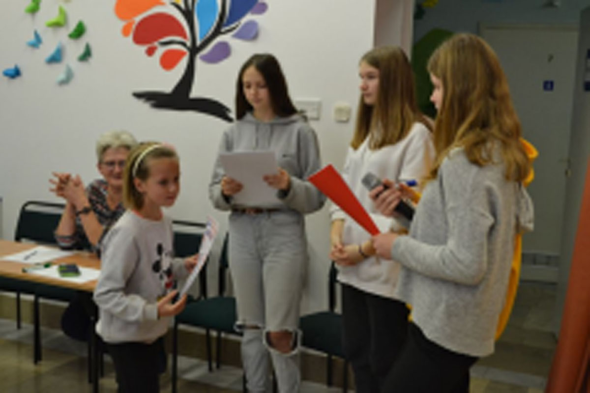 Uczniowie występują na scenie, trzymają mikrofony. Członkowie jury siedzą przy stoliku, wręczają dyplomy. Wspólne zdjęcie uczniów.