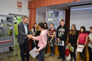 Zdjęcia przedstawiają salę, w której siedzą  uczniowie szkoły i oglądają prezentację.  Z frontu wystawione są stelaże, na których przypięte są zdjęcia przedstawiające Żołnierzy Wyklętych. Na jednym ze zdjęć przedstawiony jest dyrektor szkoły i nauczycielk