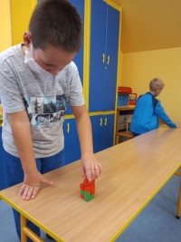 Dzieci w maseczkach siedzą na dywanie. Jedna starsza uczennica siedzi naprzeciwko dzieci i czyta im z dużej książki.