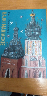 Dzieci w maseczkach siedzą na dywanie. Jedna starsza uczennica siedzi naprzeciwko dzieci i czyta im z dużej książki.