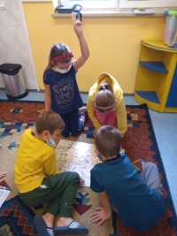 Dzieci w maseczkach siedzą na dywanie. Jedna starsza uczennica siedzi naprzeciwko dzieci i czyta im z dużej książki.