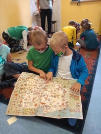 Dzieci w maseczkach siedzą na dywanie. Jedna starsza uczennica siedzi naprzeciwko dzieci i czyta im z dużej książki.