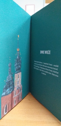 Dzieci w maseczkach siedzą na dywanie. Jedna starsza uczennica siedzi naprzeciwko dzieci i czyta im z dużej książki.
