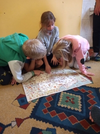 Dzieci w maseczkach siedzą na dywanie. Jedna starsza uczennica siedzi naprzeciwko dzieci i czyta im z dużej książki.