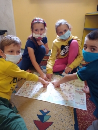 Dzieci w maseczkach siedzą na dywanie. Jedna starsza uczennica siedzi naprzeciwko dzieci i czyta im z dużej książki.