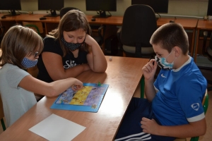 Uczniowie przy stolikach,  na których są kubki plastikowe ułożone w wieże, łyżeczki plastikowe w pojemniku i obok niego, gry planszowe i folia przeźroczysta z wynikami działań. Uczniowie siedzą na krzesłach patrzą na tablicę multimedialną.
