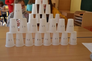 Uczniowie przy stolikach,  na których są kubki plastikowe ułożone w wieże, łyżeczki plastikowe w pojemniku i obok niego, gry planszowe i folia przeźroczysta z wynikami działań. Uczniowie siedzą na krzesłach patrzą na tablicę multimedialną.