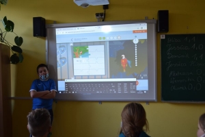 Uczniowie przy stolikach,  na których są kubki plastikowe ułożone w wieże, łyżeczki plastikowe w pojemniku i obok niego, gry planszowe i folia przeźroczysta z wynikami działań. Uczniowie siedzą na krzesłach patrzą na tablicę multimedialną.