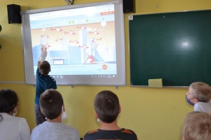 Uczniowie przy stolikach,  na których są kubki plastikowe ułożone w wieże, łyżeczki plastikowe w pojemniku i obok niego, gry planszowe i folia przeźroczysta z wynikami działań. Uczniowie siedzą na krzesłach patrzą na tablicę multimedialną.