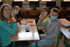 Uczniowie przy stolikach,  na których są kubki plastikowe ułożone w wieże, łyżeczki plastikowe w pojemniku i obok niego, gry planszowe i folia przeźroczysta z wynikami działań. Uczniowie siedzą na krzesłach patrzą na tablicę multimedialną.