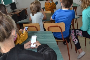 Uczniowie przy stolikach,  na których są kubki plastikowe ułożone w wieże, łyżeczki plastikowe w pojemniku i obok niego, gry planszowe i folia przeźroczysta z wynikami działań. Uczniowie siedzą na krzesłach patrzą na tablicę multimedialną.