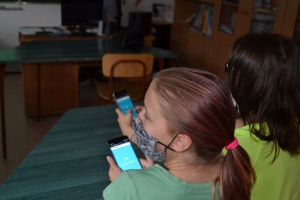 Uczniowie przy stolikach,  na których są kubki plastikowe ułożone w wieże, łyżeczki plastikowe w pojemniku i obok niego, gry planszowe i folia przeźroczysta z wynikami działań. Uczniowie siedzą na krzesłach patrzą na tablicę multimedialną.