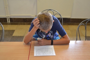 Uczniowie przy stolikach,  na których są kubki plastikowe ułożone w wieże, łyżeczki plastikowe w pojemniku i obok niego, gry planszowe i folia przeźroczysta z wynikami działań. Uczniowie siedzą na krzesłach patrzą na tablicę multimedialną.