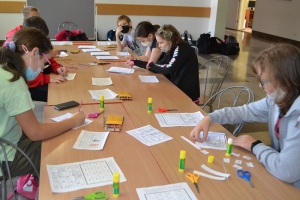 Uczniowie przy stolikach,  na których są kubki plastikowe ułożone w wieże, łyżeczki plastikowe w pojemniku i obok niego, gry planszowe i folia przeźroczysta z wynikami działań. Uczniowie siedzą na krzesłach patrzą na tablicę multimedialną.