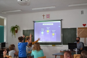 Uczniowie przy stolikach,  na których są kubki plastikowe ułożone w wieże, łyżeczki plastikowe w pojemniku i obok niego, gry planszowe i folia przeźroczysta z wynikami działań. Uczniowie siedzą na krzesłach patrzą na tablicę multimedialną.