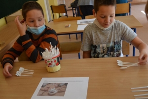 Uczniowie przy stolikach,  na których są kubki plastikowe ułożone w wieże, łyżeczki plastikowe w pojemniku i obok niego, gry planszowe i folia przeźroczysta z wynikami działań. Uczniowie siedzą na krzesłach patrzą na tablicę multimedialną.