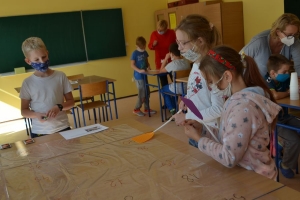 Uczniowie przy stolikach,  na których są kubki plastikowe ułożone w wieże, łyżeczki plastikowe w pojemniku i obok niego, gry planszowe i folia przeźroczysta z wynikami działań. Uczniowie siedzą na krzesłach patrzą na tablicę multimedialną.