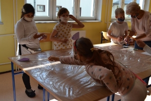 Uczniowie przy stolikach,  na których są kubki plastikowe ułożone w wieże, łyżeczki plastikowe w pojemniku i obok niego, gry planszowe i folia przeźroczysta z wynikami działań. Uczniowie siedzą na krzesłach patrzą na tablicę multimedialną.