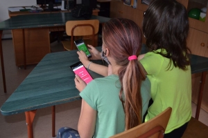 Uczniowie przy stolikach,  na których są kubki plastikowe ułożone w wieże, łyżeczki plastikowe w pojemniku i obok niego, gry planszowe i folia przeźroczysta z wynikami działań. Uczniowie siedzą na krzesłach patrzą na tablicę multimedialną.