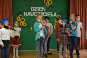 Uczniowie na scenie mówią do mikrofonu. Dyrektor mianuje pierwszoklasistów dużym ołówkiem.