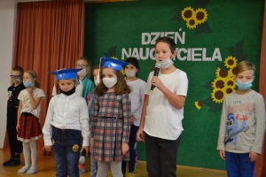 Uczniowie na scenie mówią do mikrofonu. Dyrektor mianuje pierwszoklasistów dużym ołówkiem.