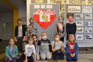 Na tle tablicy z biało-czerwonym sercem i napisem Serce dla Polski widoczna jest grupka dzieci w maseczkach. Jeden z chłopów ma uniesioną prawa rękę z palcami ułożonymi w znak zwycięstwa.SU Rabka