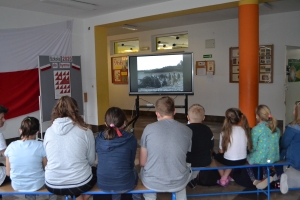 Na tle tablicy z biało-czerwonym sercem i napisem Serce dla Polski widoczna jest grupka dzieci w maseczkach. Jeden z chłopów ma uniesioną prawa rękę z palcami ułożonymi w znak zwycięstwa.ZSU Rabka