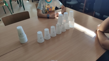 Uczniowie przy komputerach i stolikach. Układają wierze z plastikowych kubków, losują łyżeczki z zadaniami, grają w gry planszowe, rozwiązują zadania w programie komputerowym.