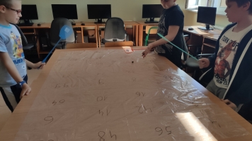 Uczniowie przy komputerach i stolikach. Układają wierze z plastikowych kubków, losują łyżeczki z zadaniami, grają w gry planszowe, rozwiązują zadania w programie komputerowym.