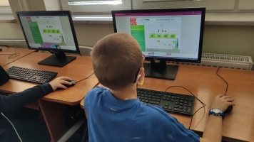 Uczniowie przy komputerach i stolikach. Układają wierze z plastikowych kubków, losują łyżeczki z zadaniami, grają w gry planszowe, rozwiązują zadania w programie komputerowym.