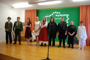 Uczniowie na scenie śpiewają pieśni. Koło sceny poczet sztandarowy, na scenie uczniowie przebrani w czarne stroje zaborców i  na biało za orła. Uczniowie składają znicze pod krzyżem.